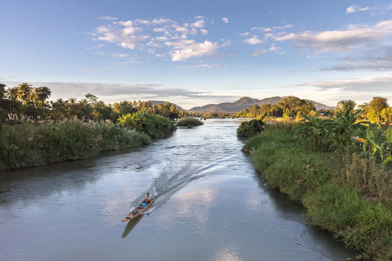 A narrow wooden boat speeding along a Mekong channel between green islands at golden hour
