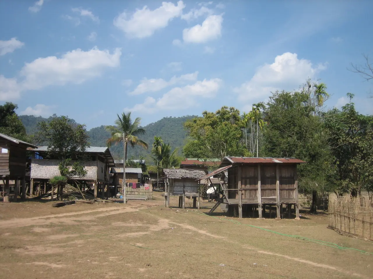 Wooden stilt houses and palm trees around a dusty open yard in a Khammouane village