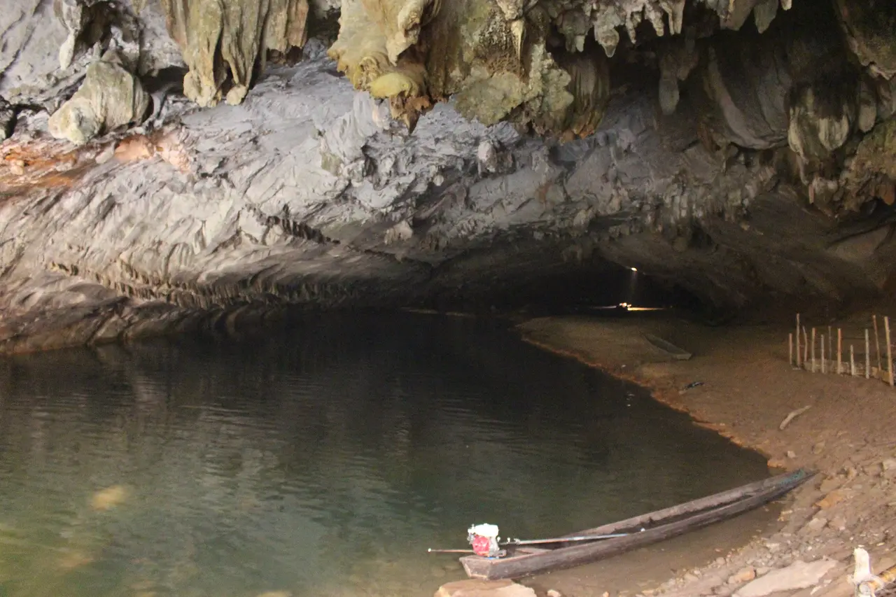 A wide, low cave mouth over still water, with a long wooden boat pulled up on a sandbank