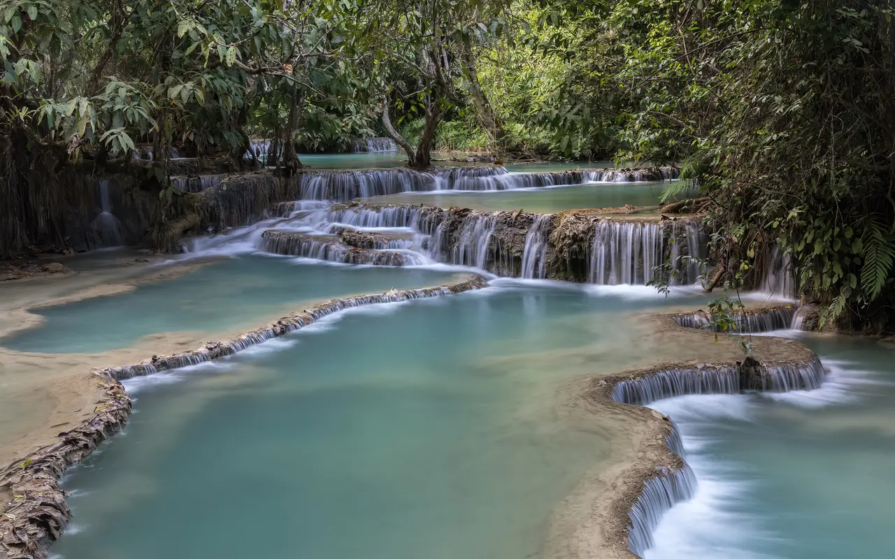 Turquoise pools stepping down over pale limestone terraces at Kuang Si Falls, surrounded by forest