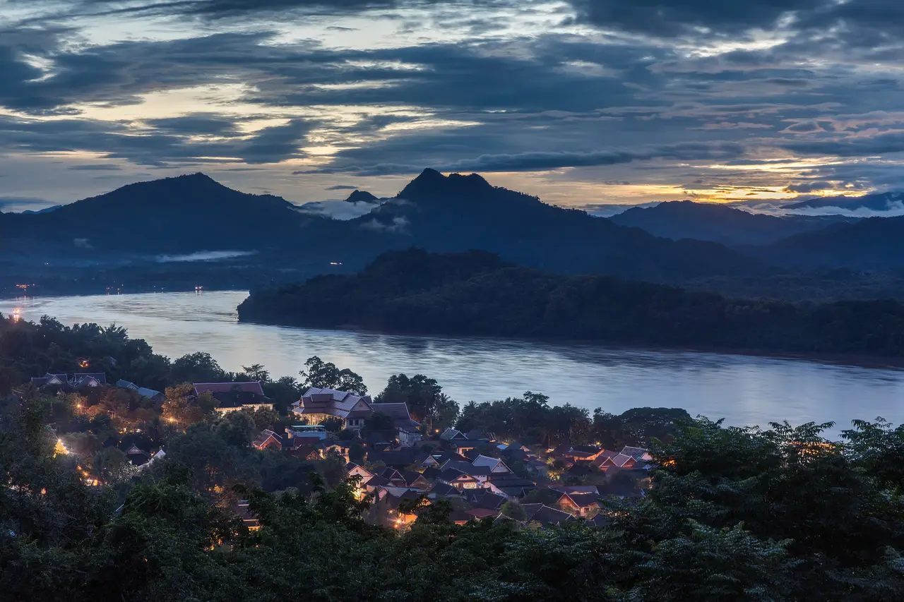Luang Prabang at dusk seen from Mount Phou Si, with lights among trees and the Mekong below layered mountains