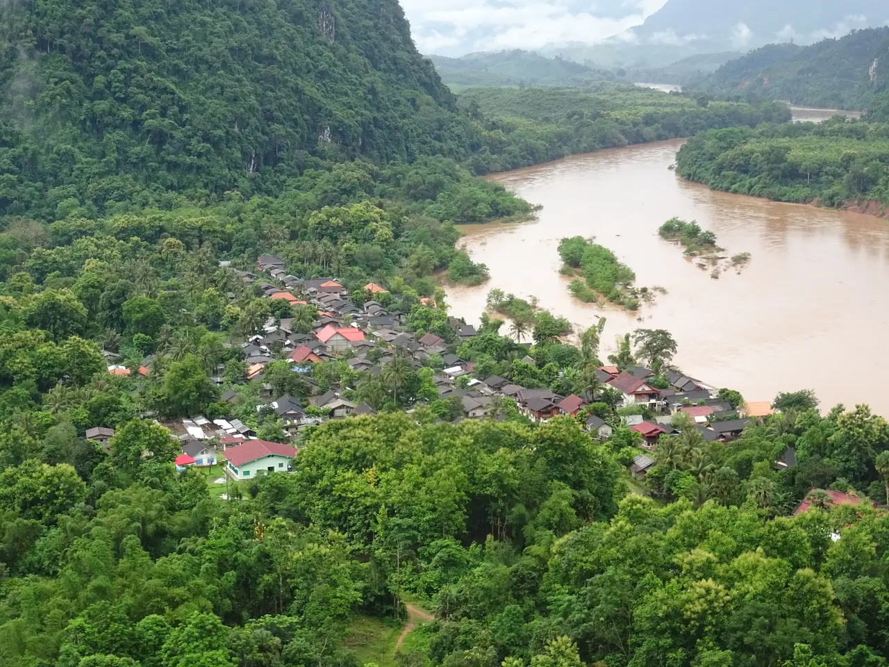 Muang Ngoi Neua seen from a hillside: a small village of roofs among trees beside the broad Nam Ou