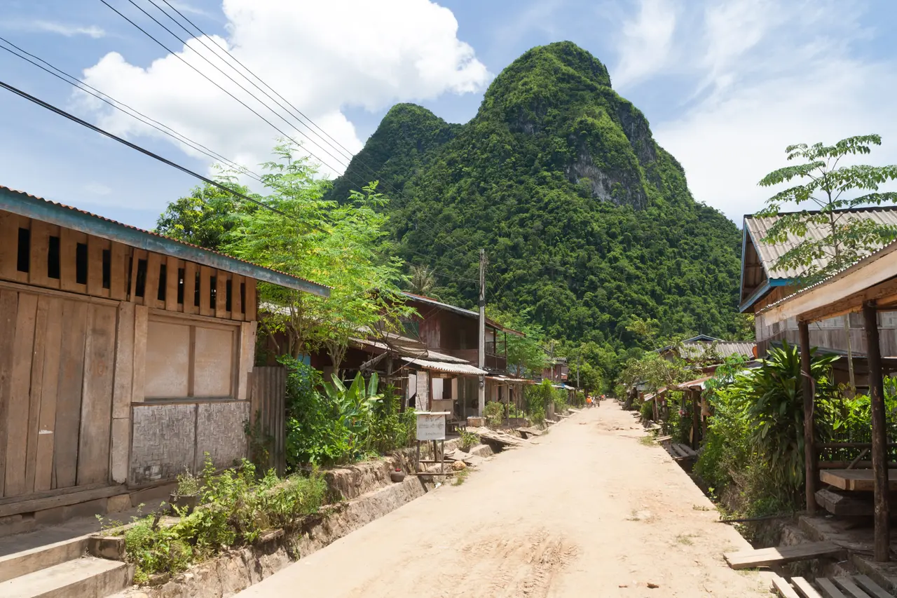 A sandy lane between wooden houses in Muang Ngoi Neua, with a steep green peak behind