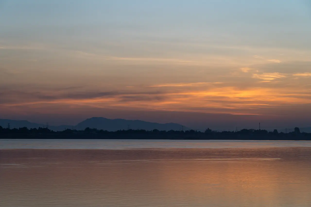 An orange sunset over the wide, calm Mekong at Pakse with low hills on the far bank