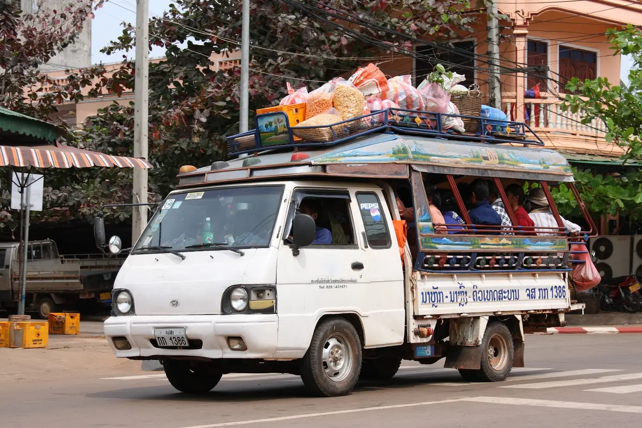 A white pickup converted into a songthaew, with bench seats in the back and luggage piled on the roof
