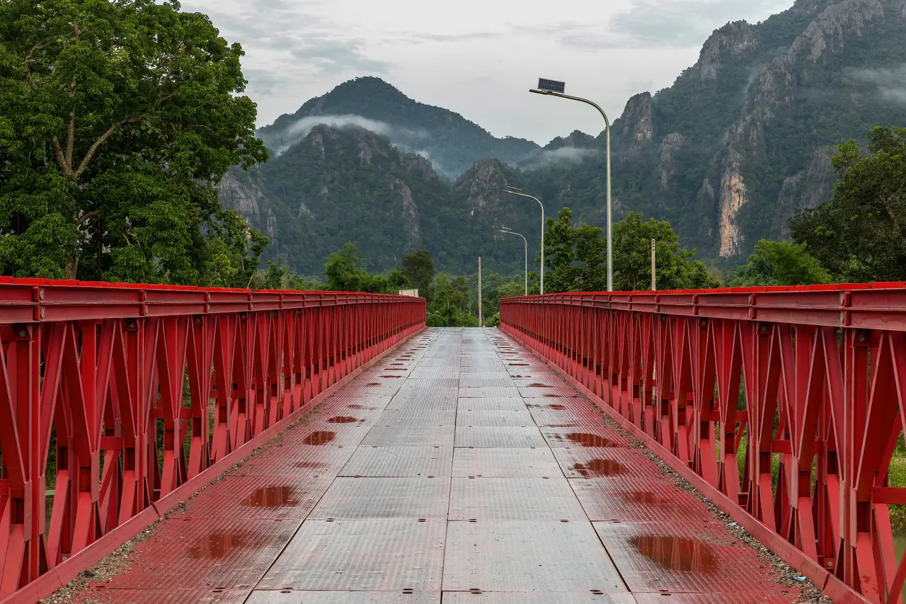 A red metal footbridge over the Nam Song leading towards misty karst mountains