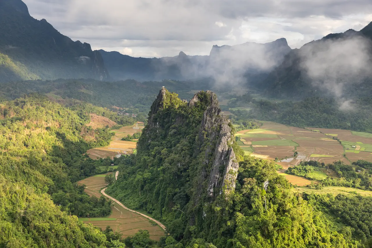 Green karst peaks and a winding road through rice fields below, seen from a mountain top near Vang Vieng