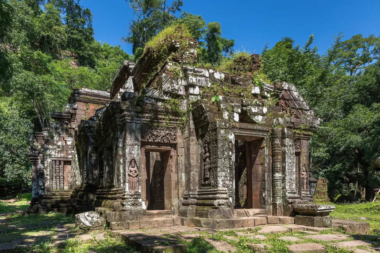 A ruined sandstone Khmer temple sanctuary at Vat Phou, overgrown with plants under a blue sky